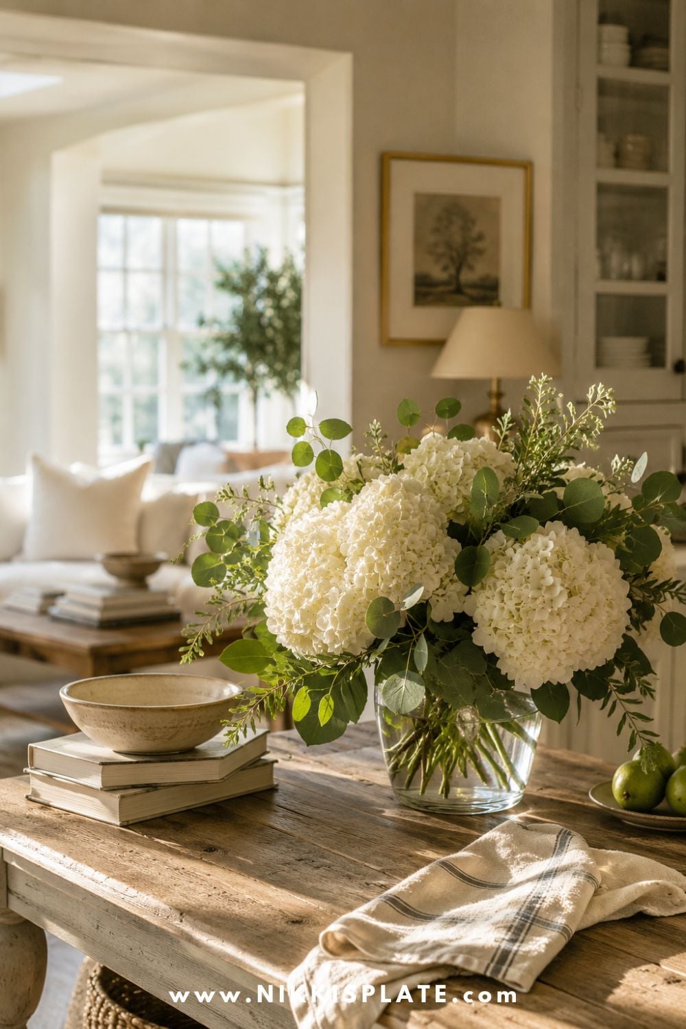 Nancy Meyers aesthetic home decor with fresh white hydrangeas and greenery in a glass vase, rustic wood table, warm neutral living room, vintage books, linen towel, and cozy classic cottage style.