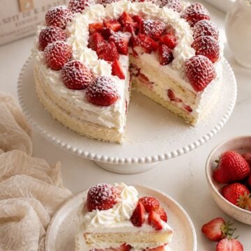 Homemade strawberry shortcake with a slice served on a plate, topped with fresh strawberries, powdered sugar, and homemade whipped cream.