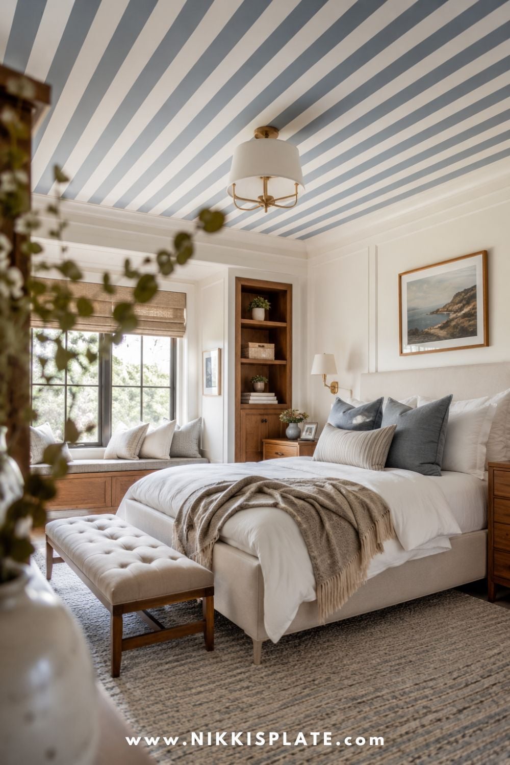 Bright bedroom with a blue-gray striped ceiling, white upholstered bed, cozy window seat, warm wood accents, and layered neutral bedding.