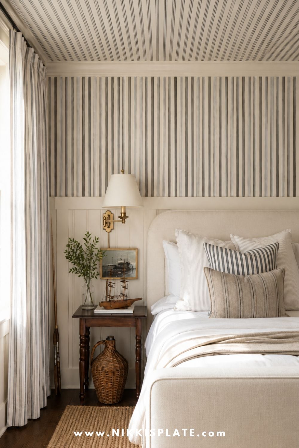 Cozy bedroom with striped wallpaper extending onto the ceiling, a neutral upholstered bed, layered pillows, and classic coastal decor.
