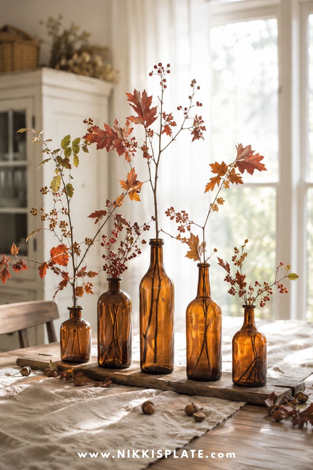 Amber glass bottles filled with airy burgundy, rust and olive autumn branches arranged along a farmhouse dining table.