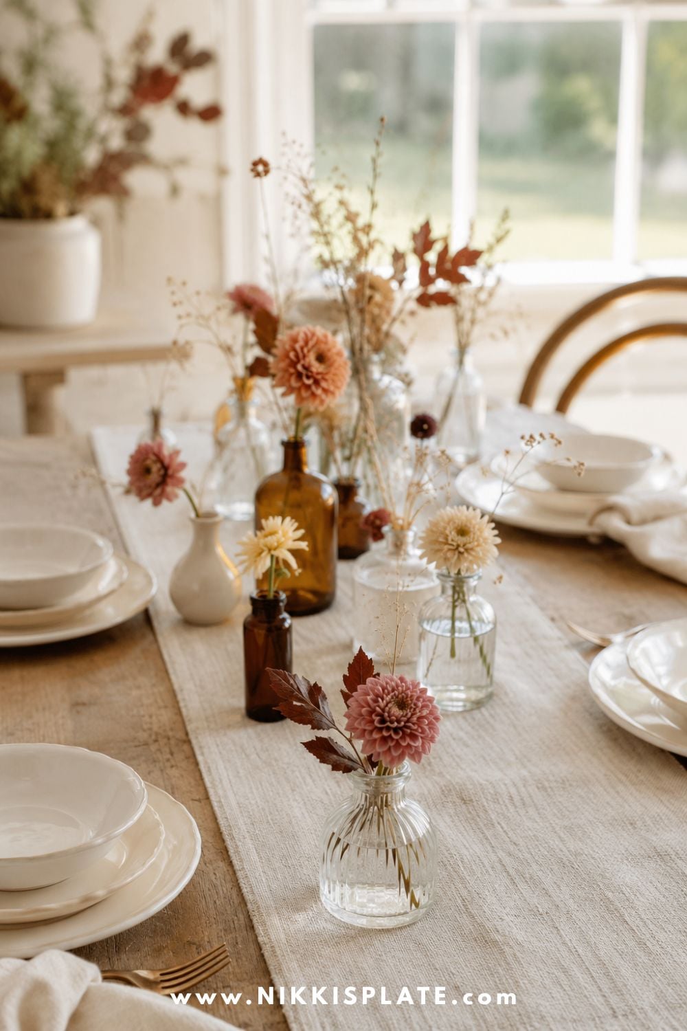 Small amber and clear bud vases filled with dahlias, chrysanthemums and autumn branches along a farmhouse table.