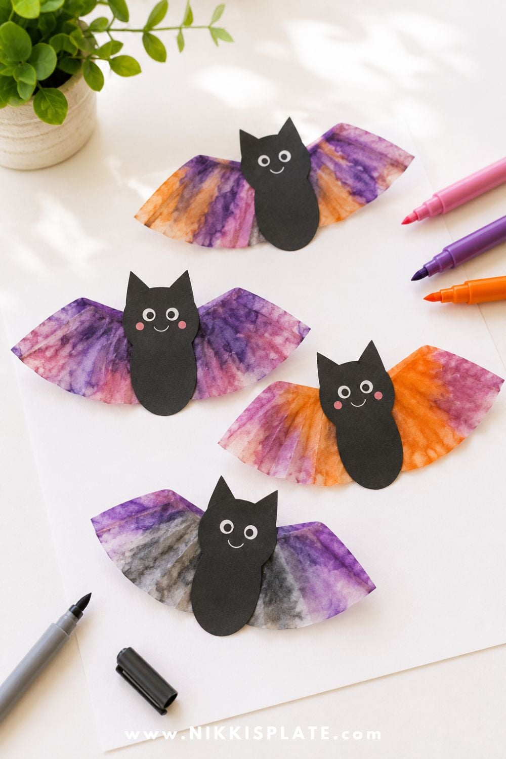 Coffee filter bat craft with black paper bat body and colorful marker-dyed wings, shown on a bright craft table for an easy Halloween craft for kids.