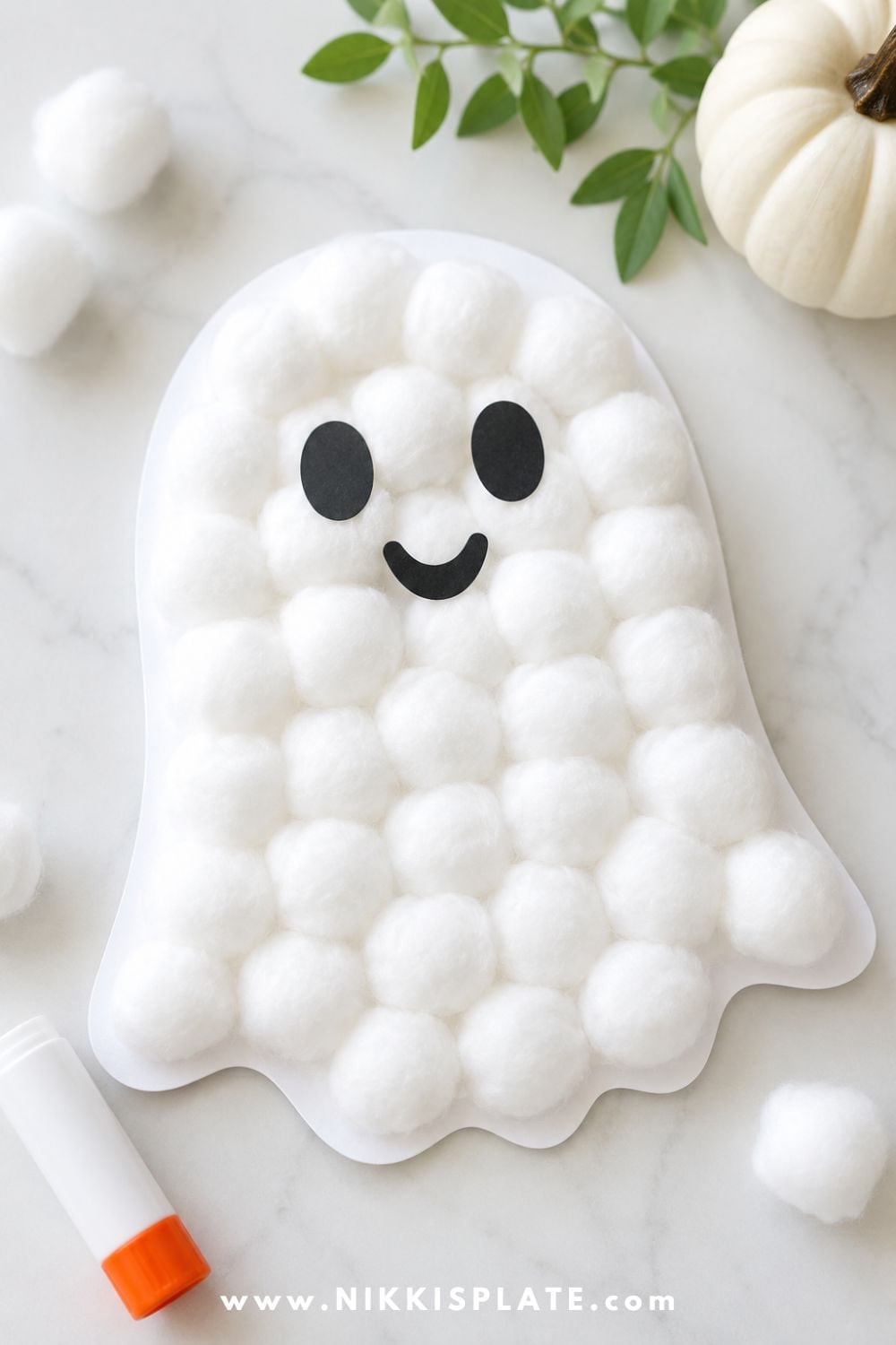 Fluffy cotton ball ghost craft on white paper with a cute ghost face, styled on a clean craft table for a simple and low-mess Halloween craft for kids.