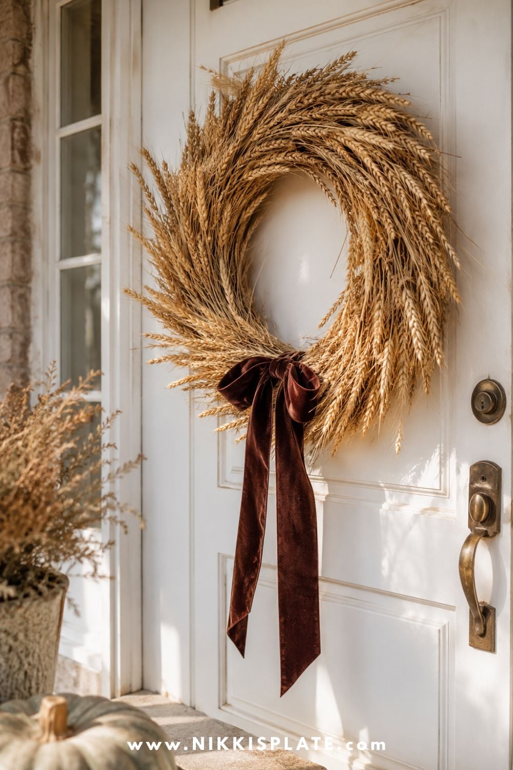 Handmade dried wheat wreath finished with a deep burgundy velvet ribbon and displayed on a warm white farmhouse door.