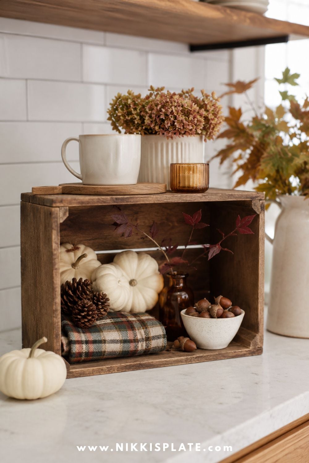 Small stained wood crate filled with cream pumpkins, pinecones, amber glass, plaid fabric and acorns on a kitchen counter.