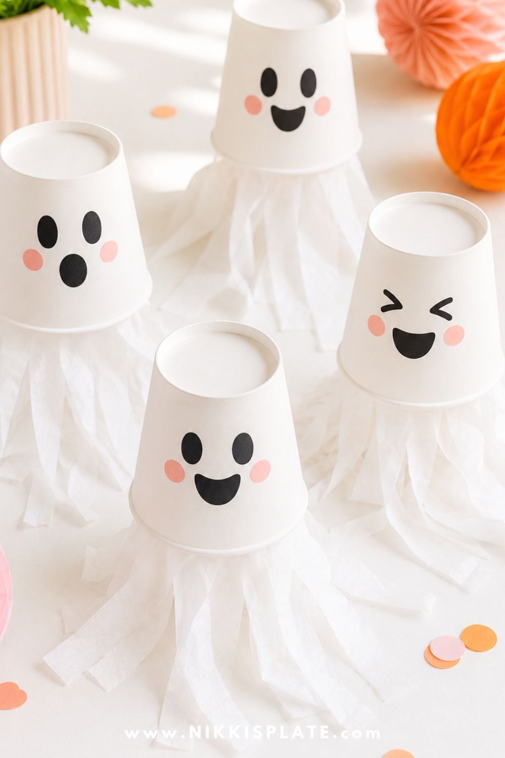 White paper cup ghost crafts with friendly faces and tissue paper streamers on a clean white table for an easy Halloween party craft for kids.