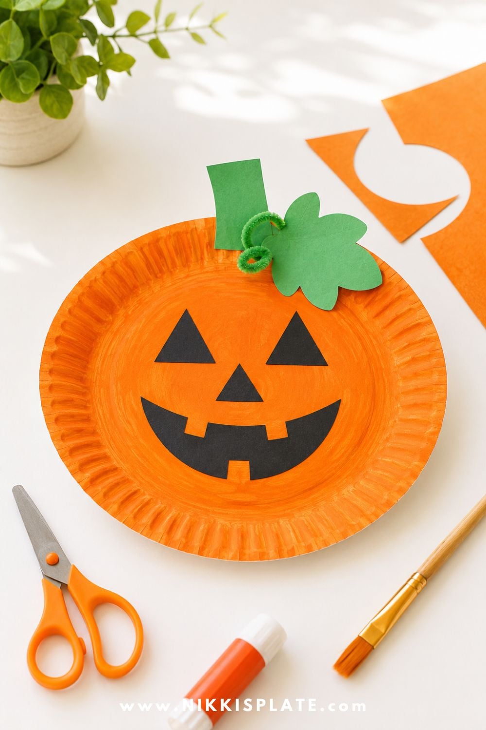 Orange paper plate pumpkin craft with a friendly jack-o-lantern face, green paper stem, and simple craft supplies on a bright white table for an easy Halloween craft for kids.