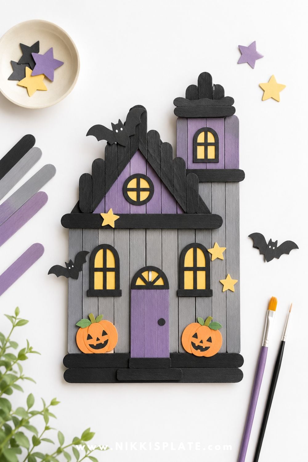 Handmade popsicle stick haunted house craft decorated with paper pumpkins, bats, stars, and windows for a cute Halloween craft activity for kids.