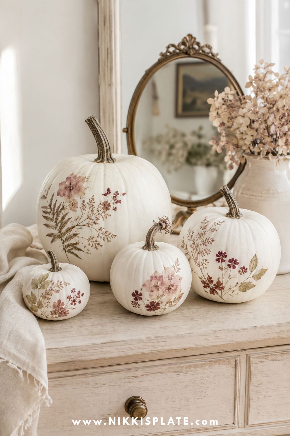 White faux pumpkins decorated with pressed blush flowers, burgundy botanicals and muted green leaves on a pale wood dresser.