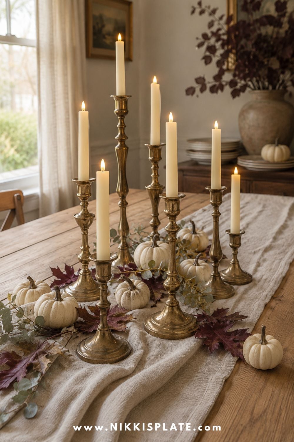 Mismatched thrifted brass candlesticks with ivory tapers, miniature pumpkins and dried leaves on a linen-covered fall table.
