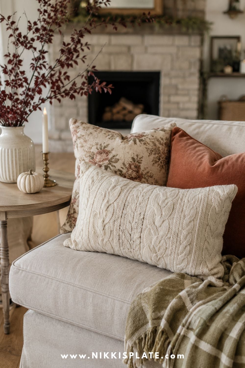Upcycled cream cable-knit sweater pillow styled with rust velvet, faded floral fabric and a cozy olive throw for fall.