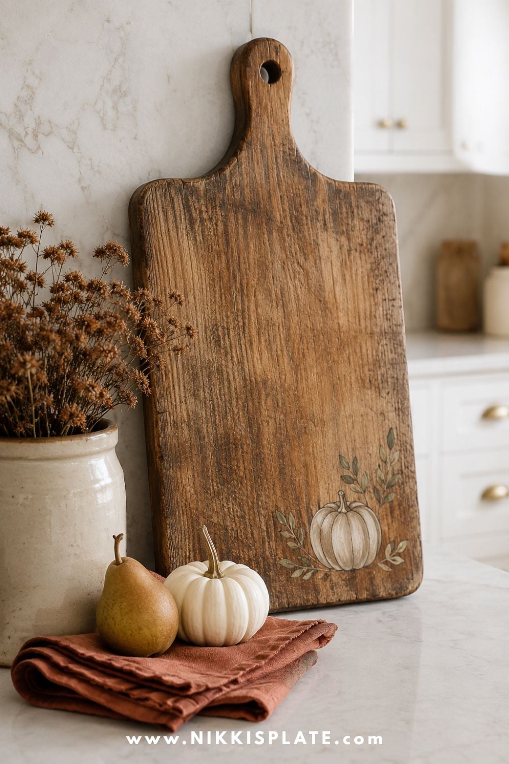 Weathered wood cutting board decorated with a painted cream pumpkin and olive leaves beside simple farmhouse kitchen accents.