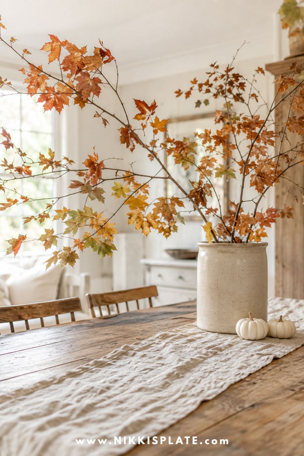 Autumn branches with faded rust and golden leaves arranged in a vintage cream vase on a farmhouse dining table.