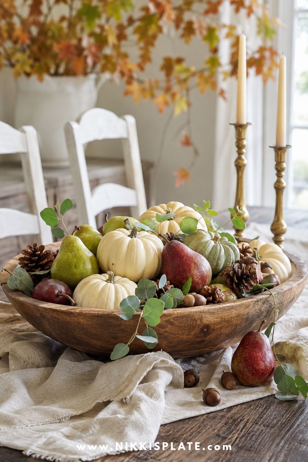 Vintage wooden bowl filled with neutral pumpkins, pears, pinecones, acorns and eucalyptus on a farmhouse dining table.