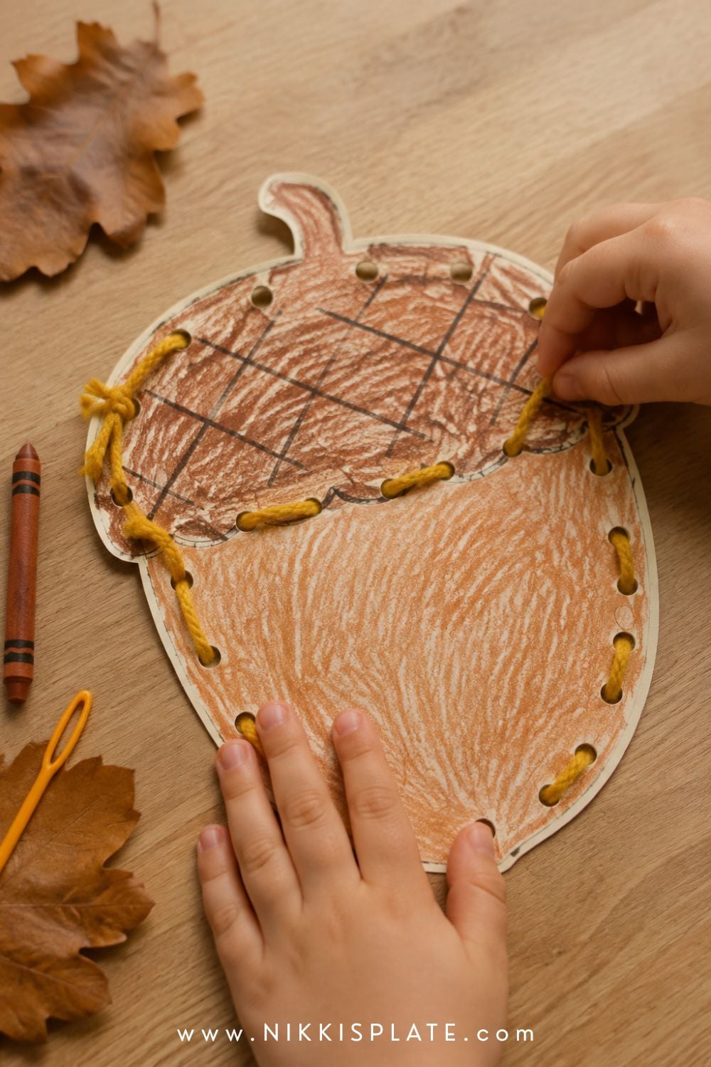 Child threading mustard yarn through a printable acorn lacing card coloured with brown crayon.