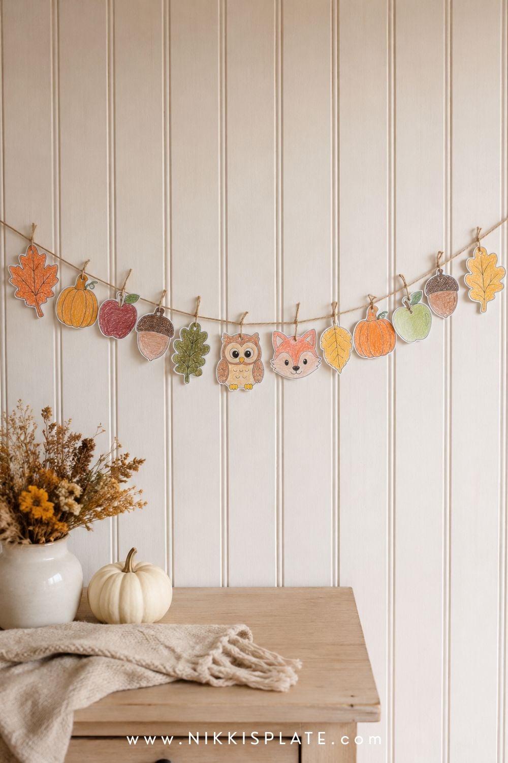 Child-made autumn paper garland with leaves, pumpkins, apples, acorns and woodland animals hanging on a white wall.