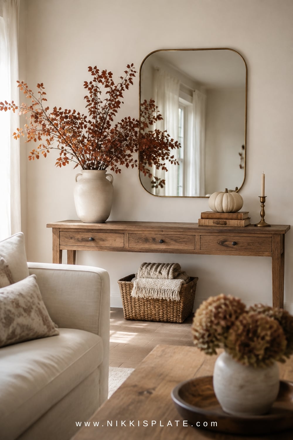 Oak console styled with a brass mirror, tall rust branches, vintage books and a cream pumpkin on a blank wall.