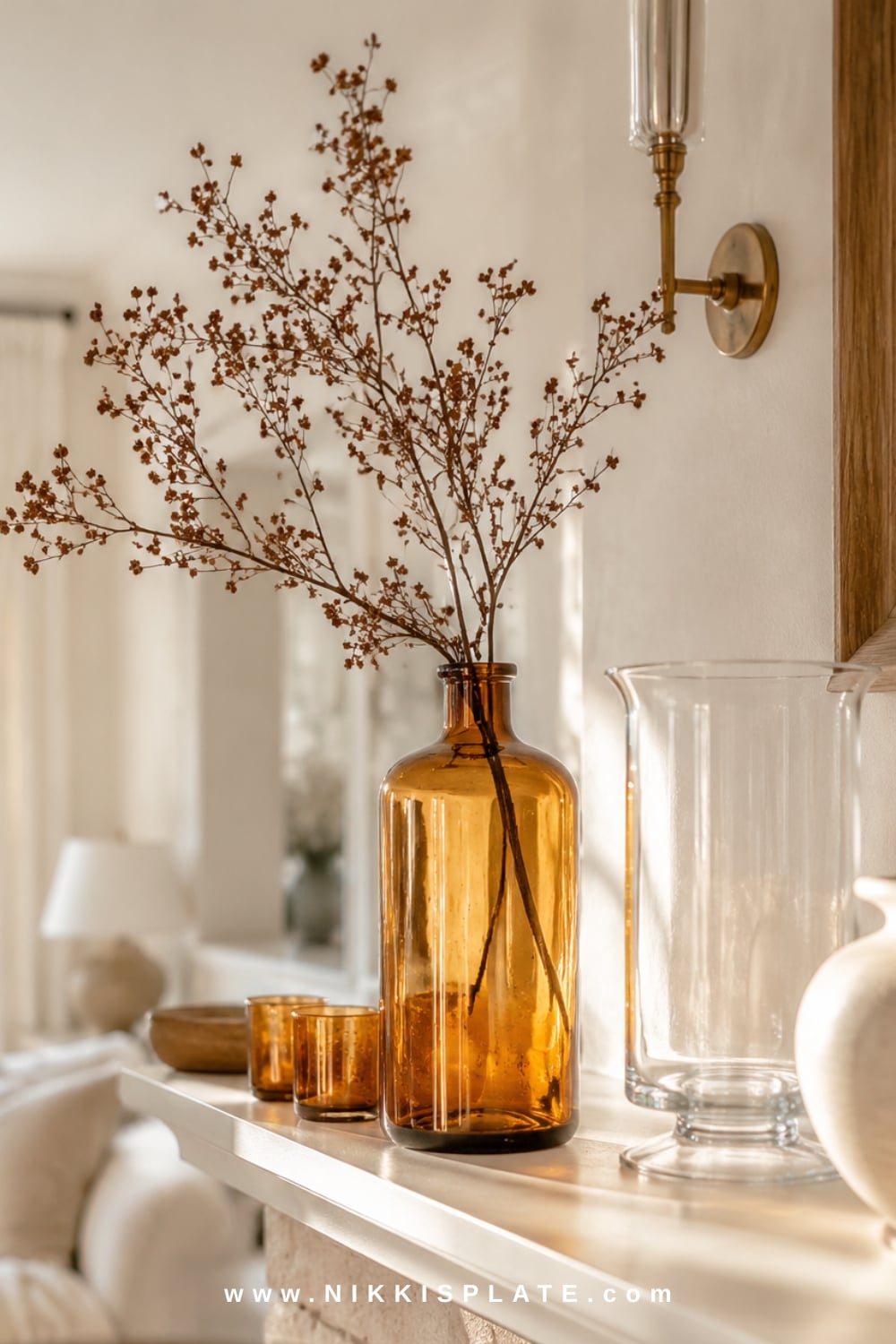 Amber glass fall mantel decor with vintage bottles, candleholders, cream pottery and soft autumn branches glowing in natural light.