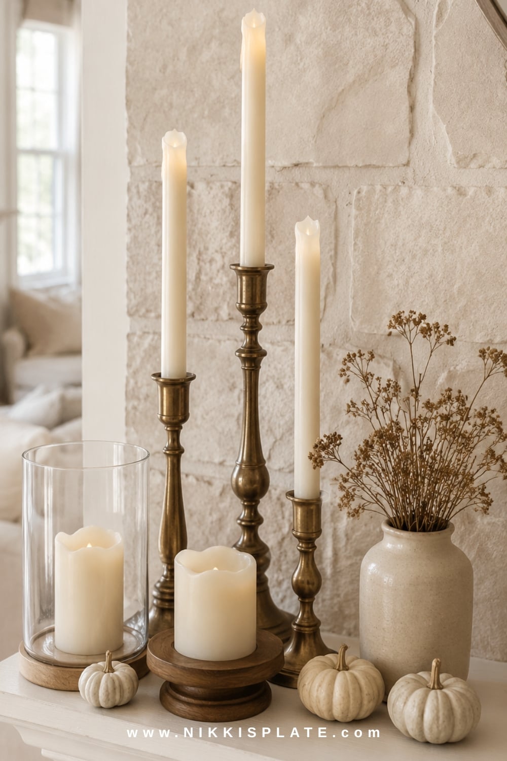 Mixed brass and warm wood candlesticks with ivory tapers, cream pumpkins and muted foliage on a cozy fall mantel.
