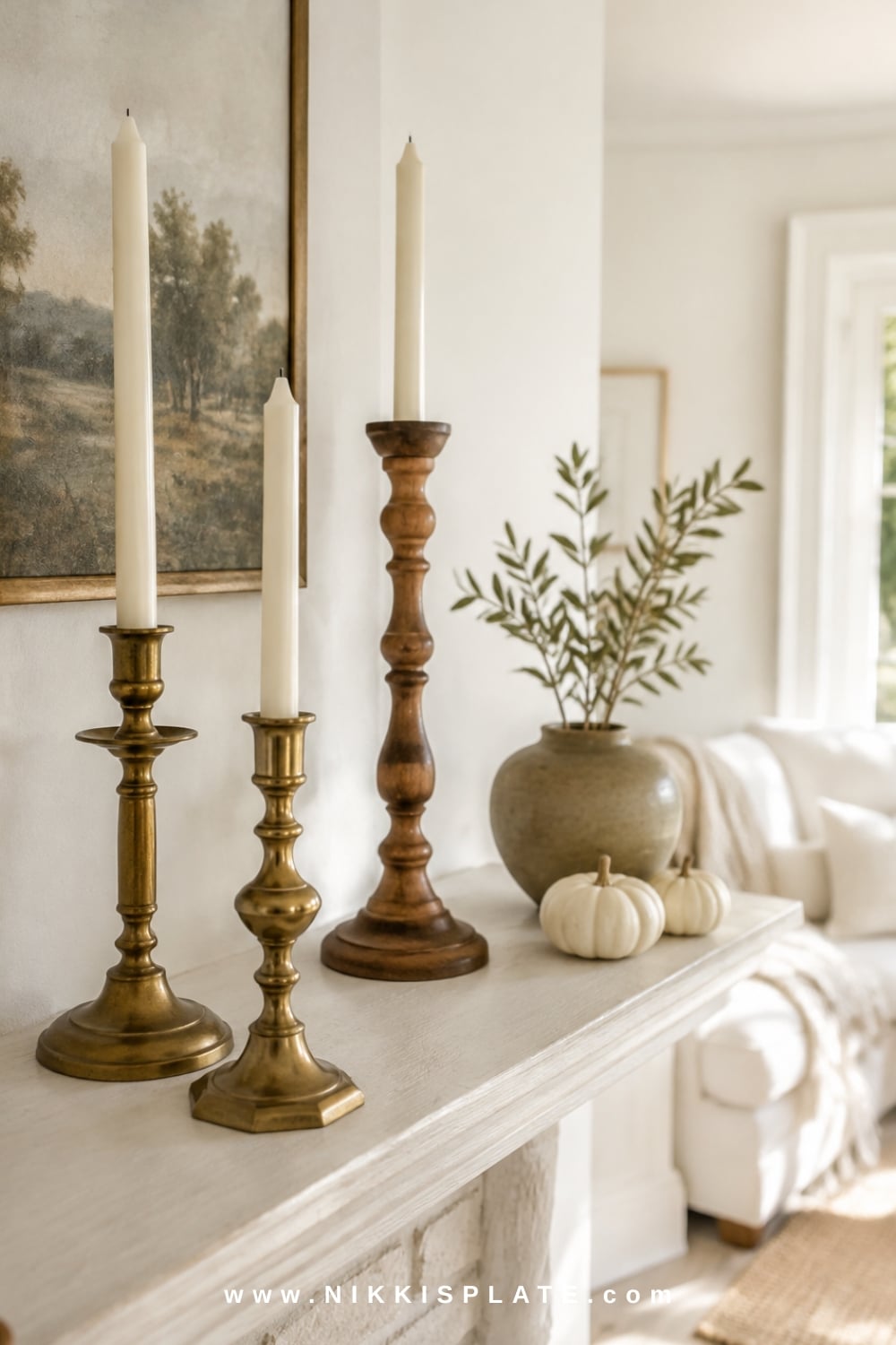 Tiered flameless candle cluster with brass, wood and glass holders beside pale pumpkins and dried stems on a farmhouse mantel.