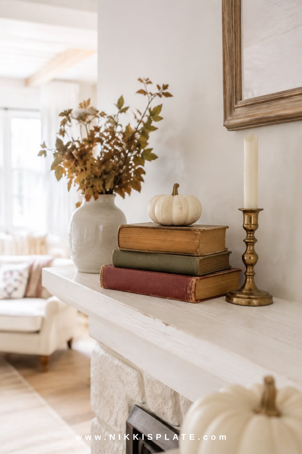 Vintage books stacked for height beside a cream pumpkin, brass candleholder and loose fall branches on a white mantel.