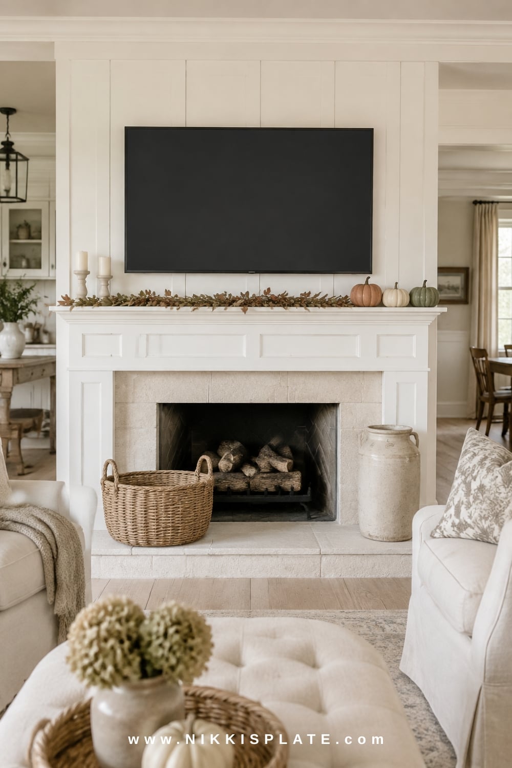 Low-profile fall mantel decor beneath a television with muted foliage, cream pumpkins and short candleholders on a white fireplace.
