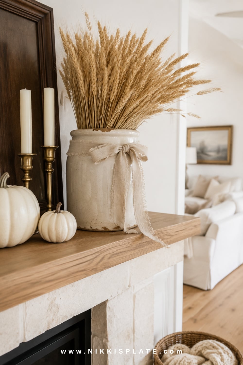 Vintage cream crock filled with wheat stems beside white pumpkins and brass candlesticks on a warm farmhouse mantel.