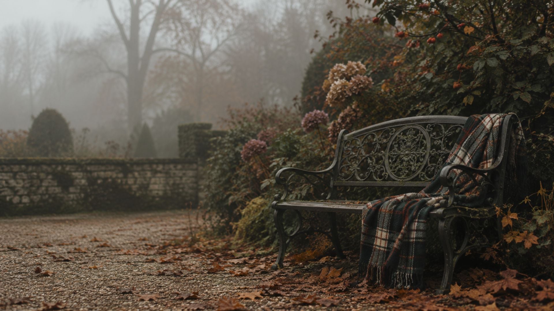 Moody fall desktop wallpaper featuring an ornate garden bench, plaid blanket, fallen leaves and faded hydrangeas in the fog.