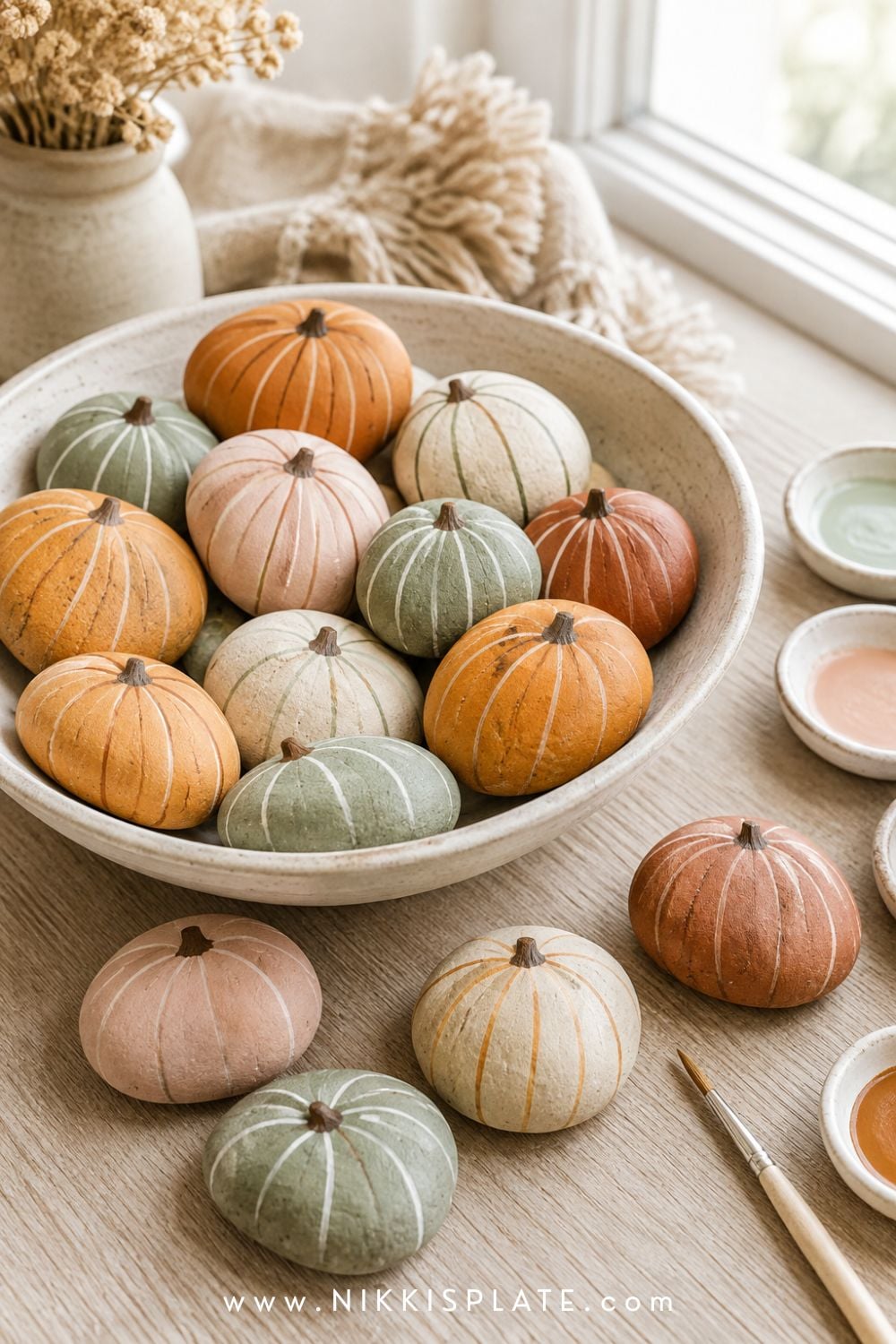 Painted pumpkin rocks in muted orange, cream, blush and green displayed in a shallow ceramic bowl.