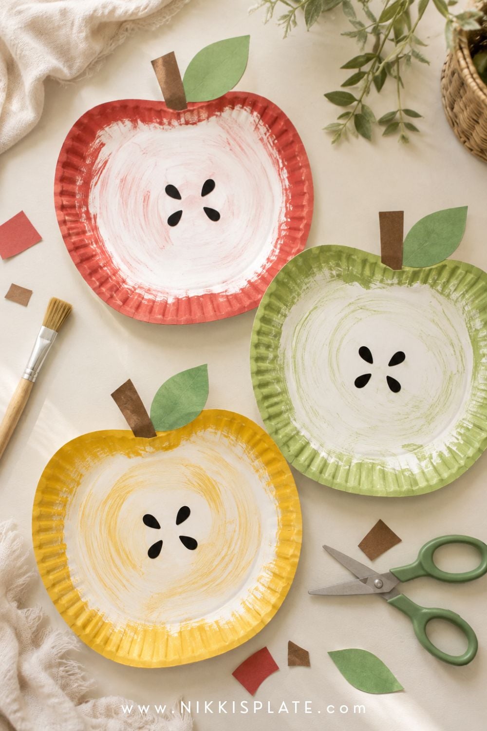 Paper plate apple crafts for kids painted red, green and yellow with brown stems, leaves and paper seeds.