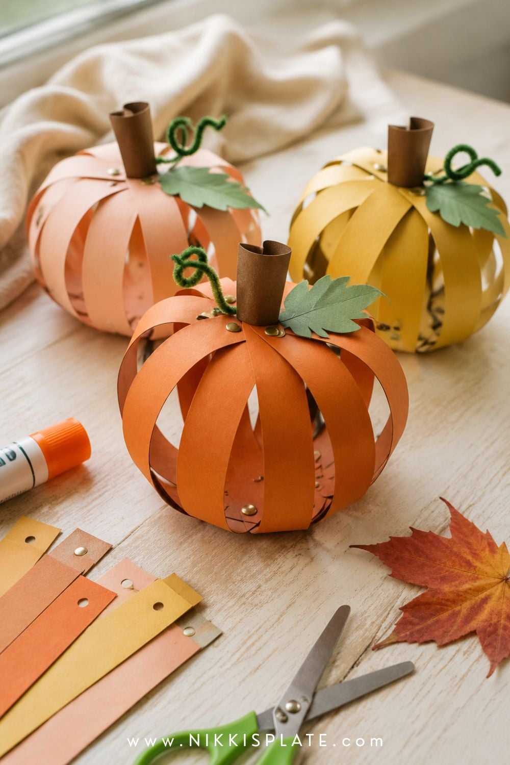 Three child-made paper strip pumpkins in rust, blush and ochre cardstock with paper leaves and curly vines.