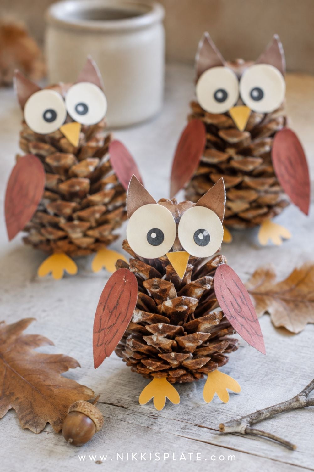 Three child-made pinecone owl crafts with paper eyes, wings and beaks displayed among autumn leaves.