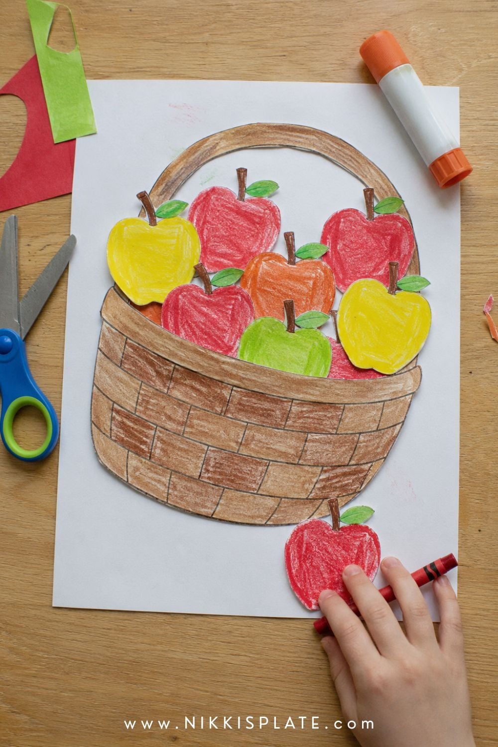 Printable apple basket craft filled with child-coloured red, yellow and green paper apples beside crayons and scissors.