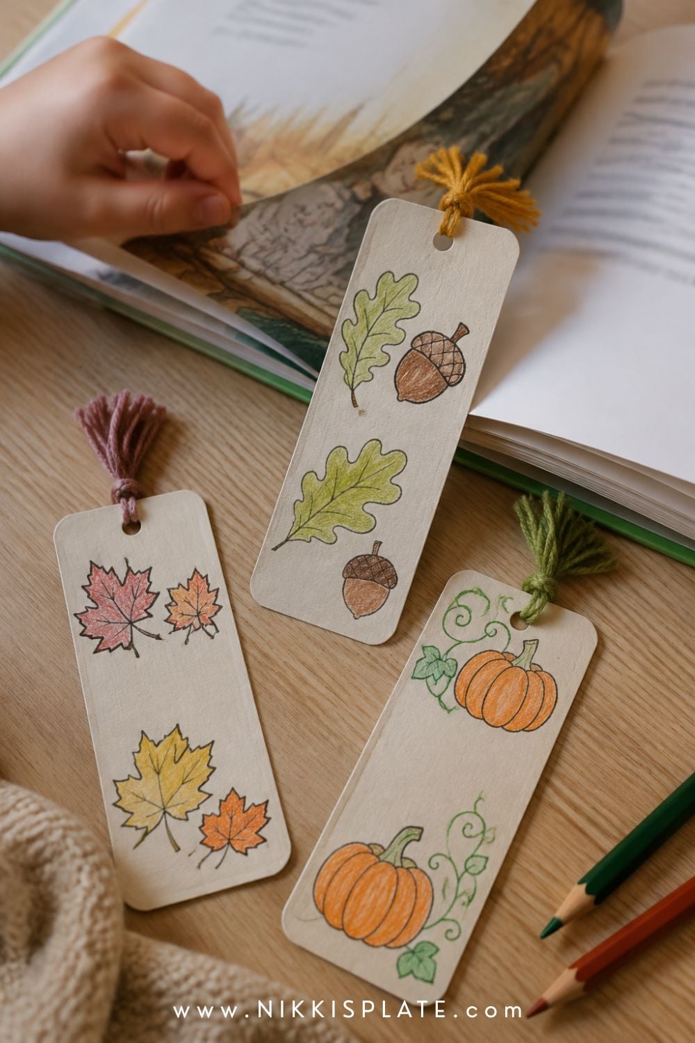 Three child-coloured printable fall bookmarks with leaves, acorns, pumpkins and yarn tassels beside an open book.