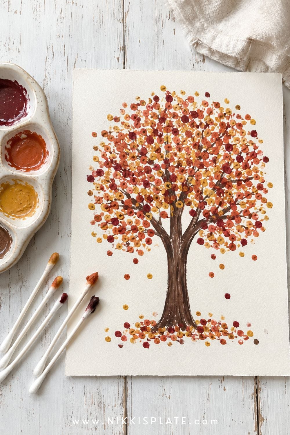 Q-tip painted autumn tree with dotted rust, orange and yellow leaves on white paper beside washable paints.