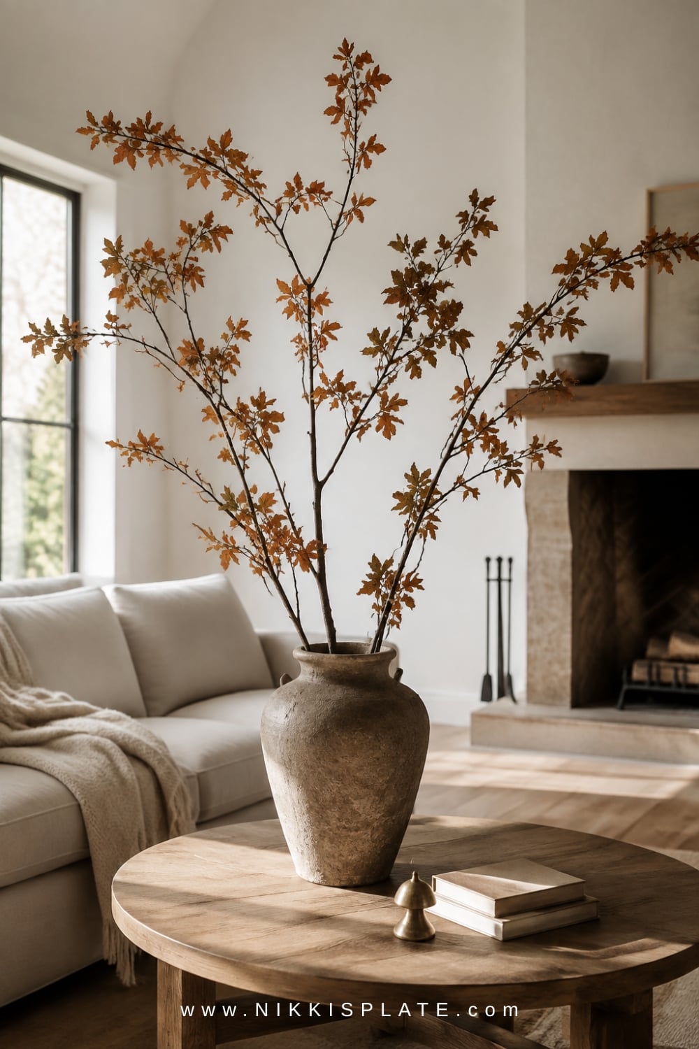 Oversized cream vase filled with tall rust, olive and brown fall branches on a round oak coffee table.