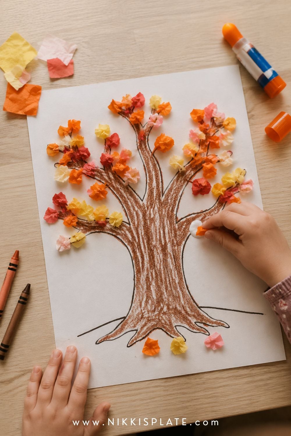 Child pressing orange tissue paper onto a printable fall tree craft surrounded by crayons, glue and autumn-coloured paper.