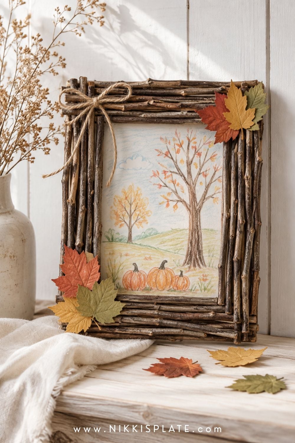Handmade twig picture frame decorated with autumn leaves and displaying a simple fall landscape drawing.