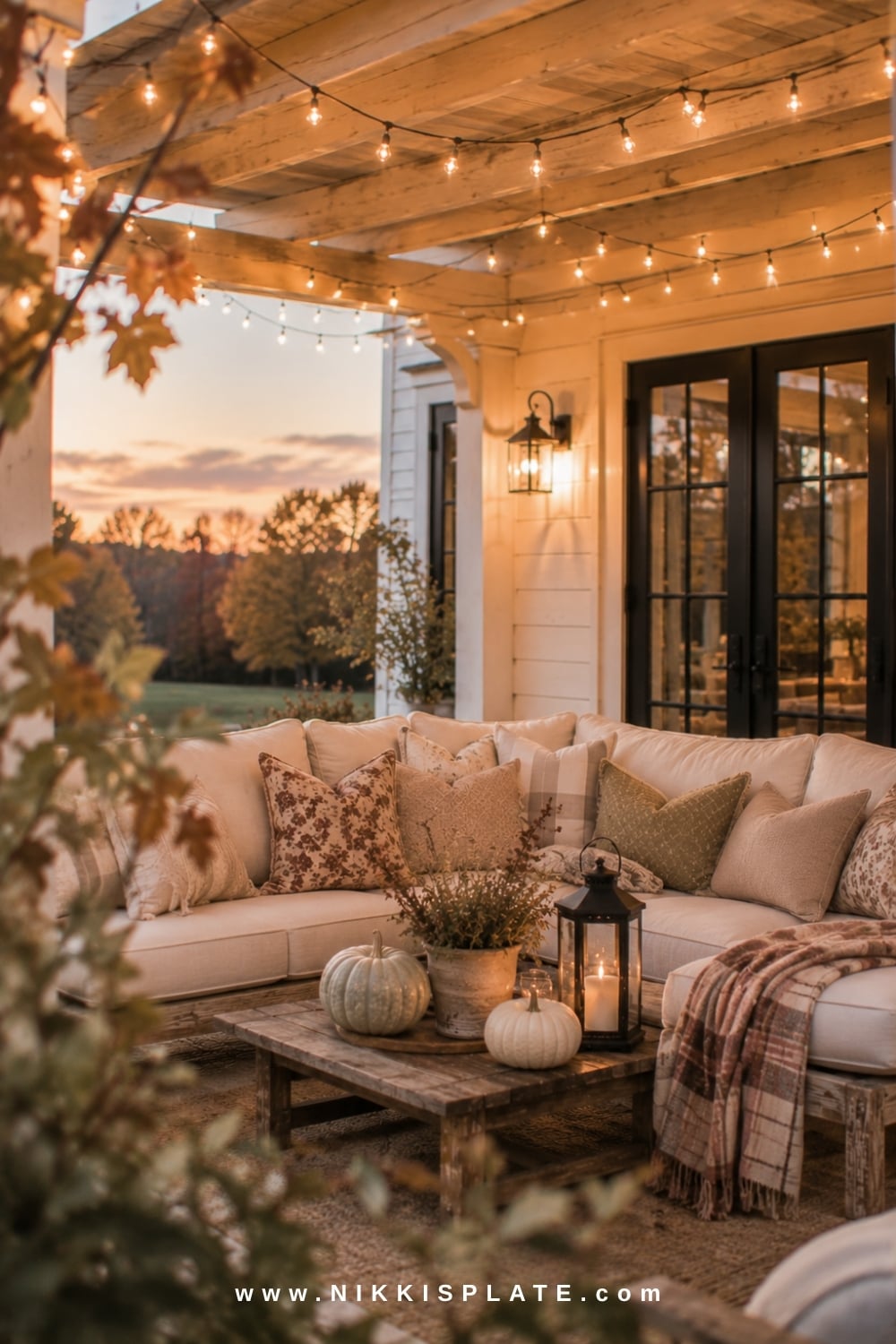Covered farmhouse patio with warm string lights, neutral seating, plaid blankets and pale pumpkins.