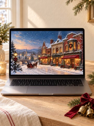 Laptop displaying a vintage Christmas village wallpaper on a wood desk with brass bells and a red velvet bow.