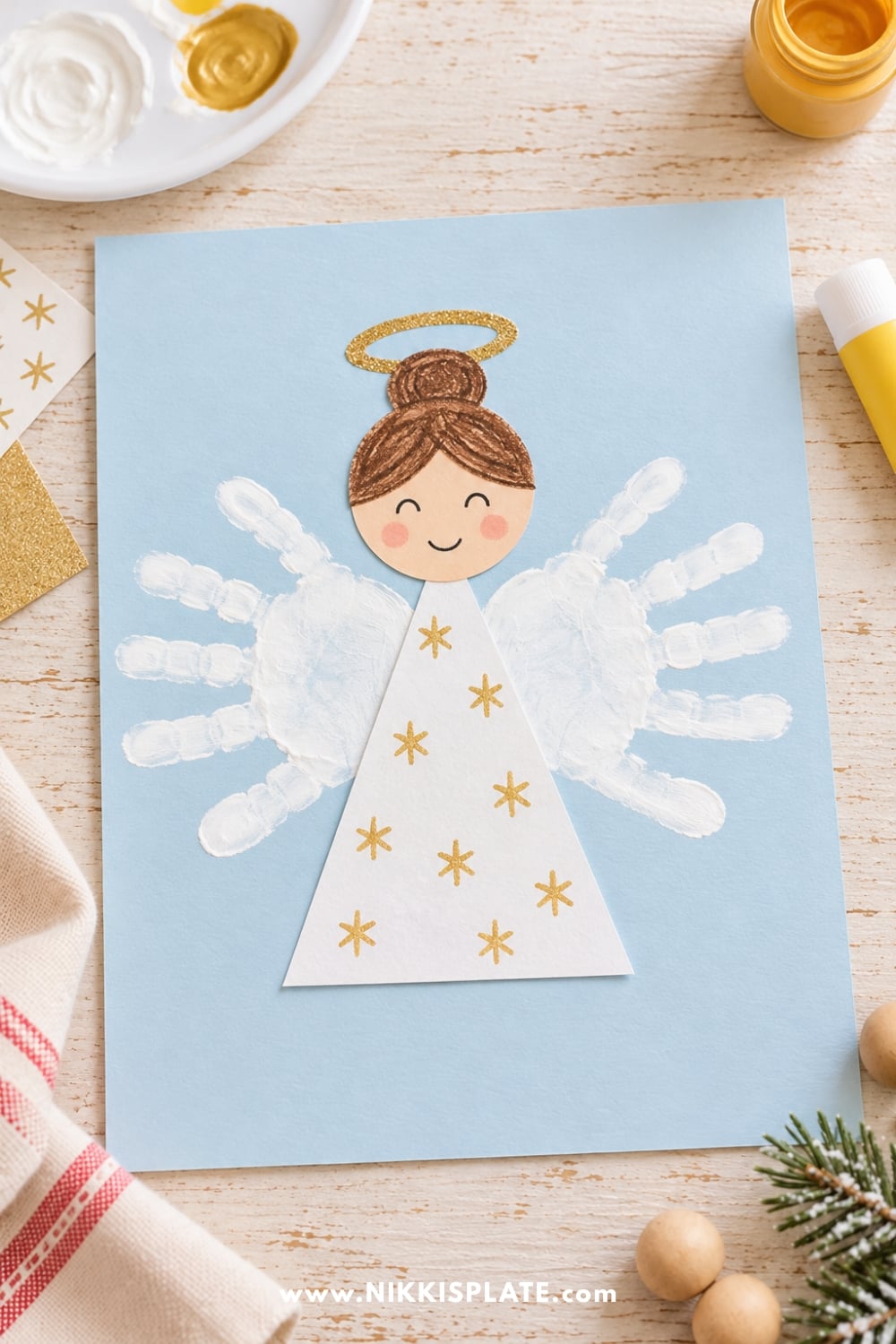 Easy Christmas angel craft for kids featuring white handprint wings, a paper gown, smiling face, and golden halo.