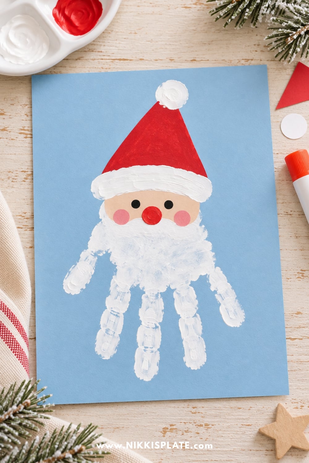 Santa handprint Christmas craft with a white painted handprint beard, red Santa hat, and rosy cheeks on blue cardstock.